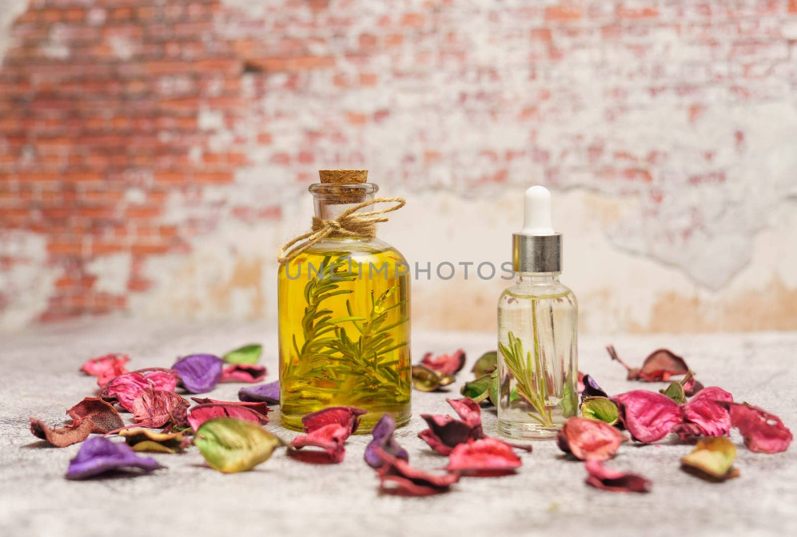 bottle and dropper with rosemary oil with fresh branches inside surrounded by colorful flower leaves and a brick wall in the background