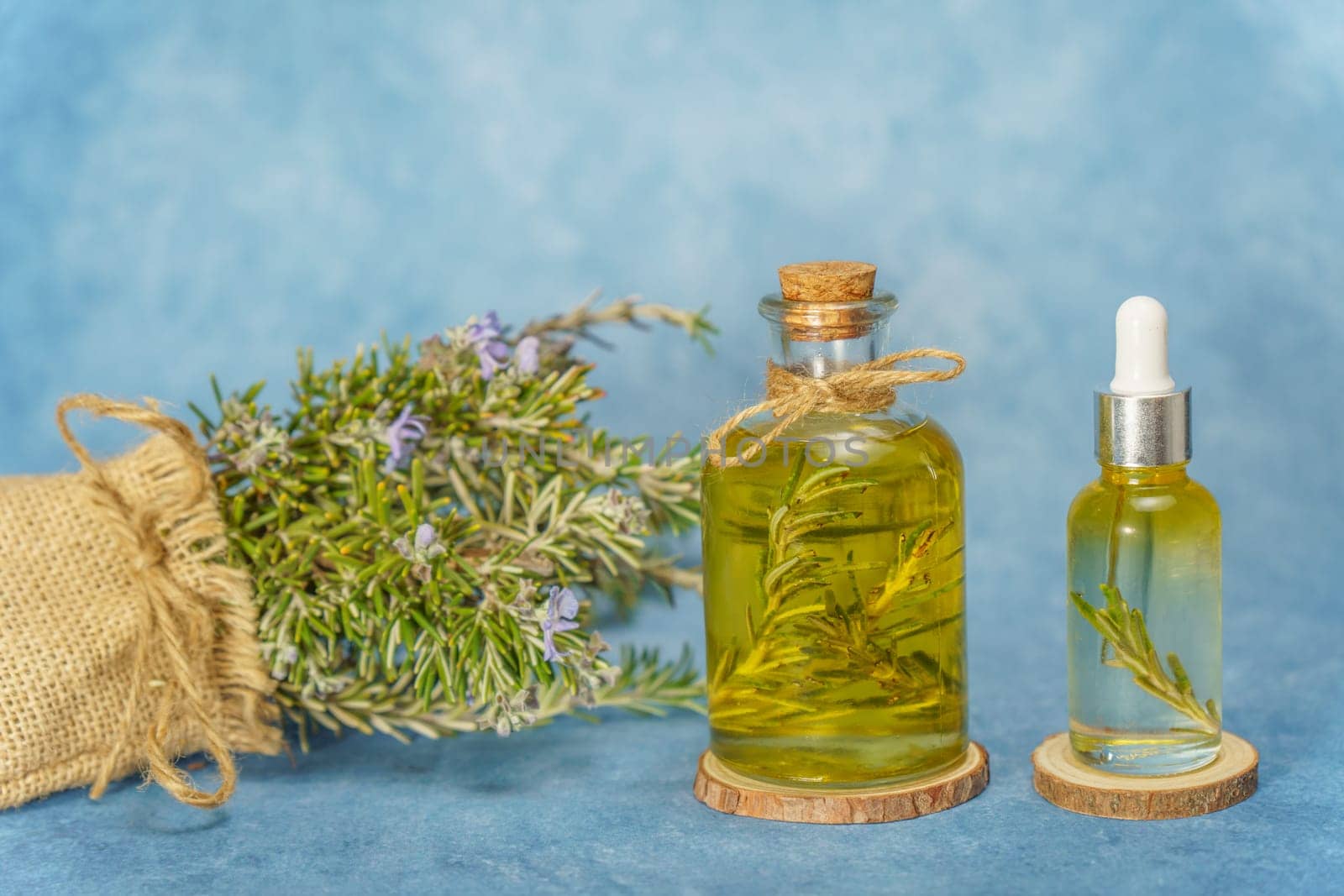 glass bottle and dropper with rosemary essential oil with fresh branches inside next to a raffia bag with freshly cut flowering rosemary branches on a blue background
