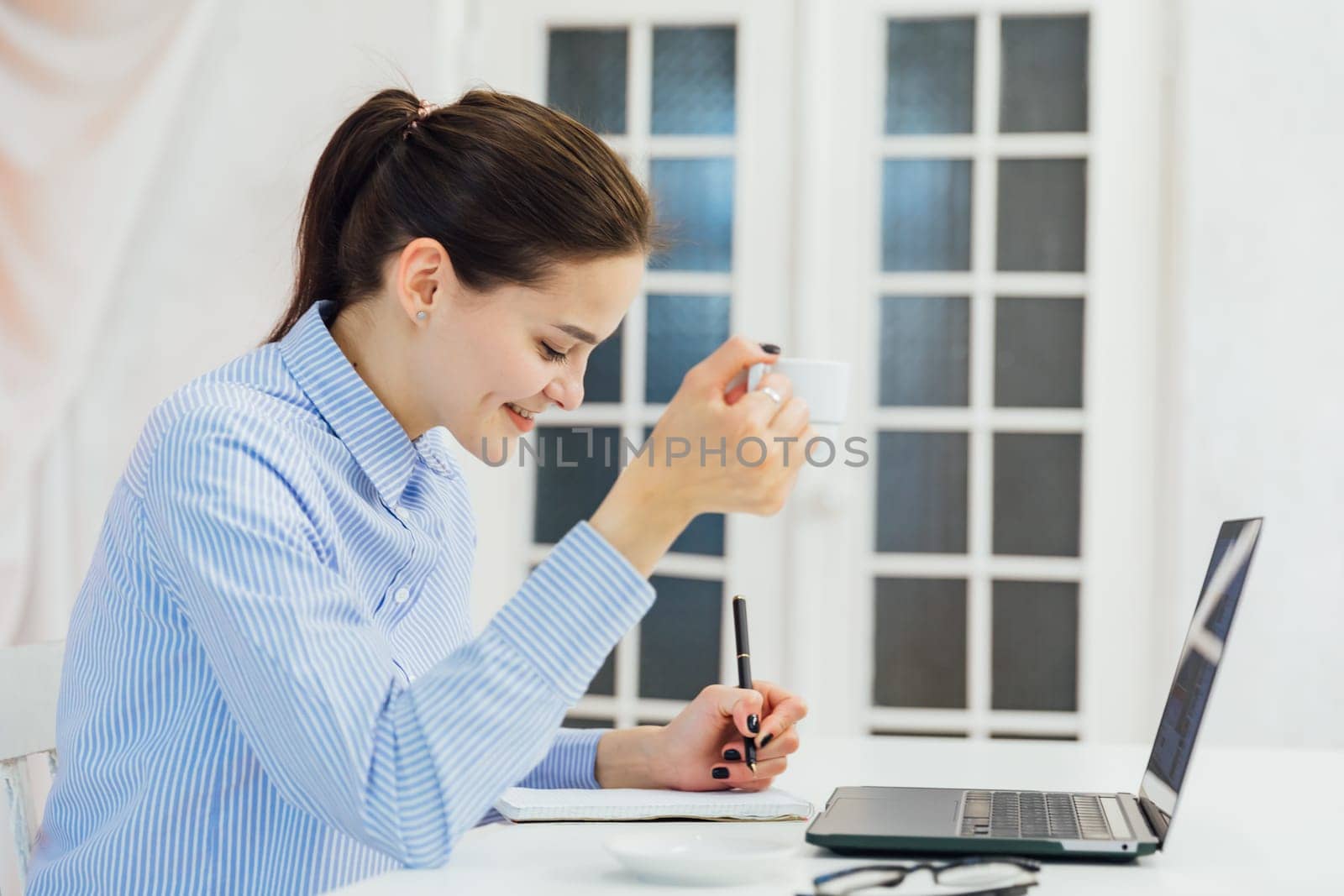 a woman at a computer with a mug writes in a notebook in the office at a remote work Internet communication IT work by Simakov