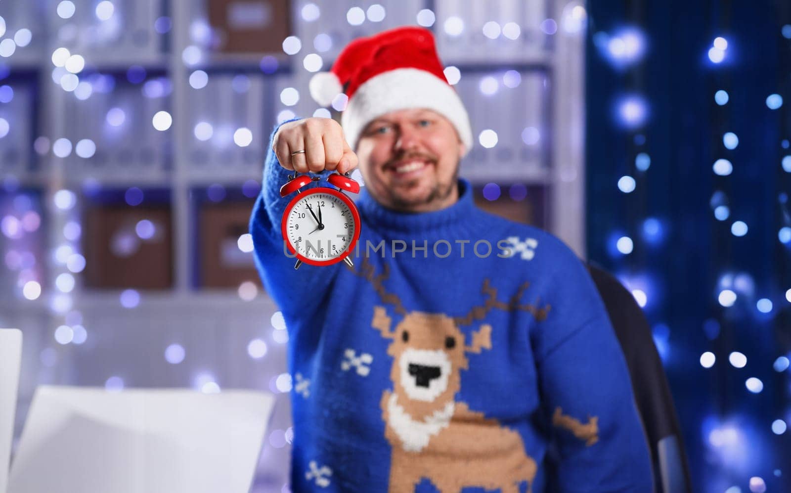 Bearded smiling man wearing warm blue sweater with traditional deer hold arm red alarm clock. Within inch coming 2024 year with glowing garland background concept closeup