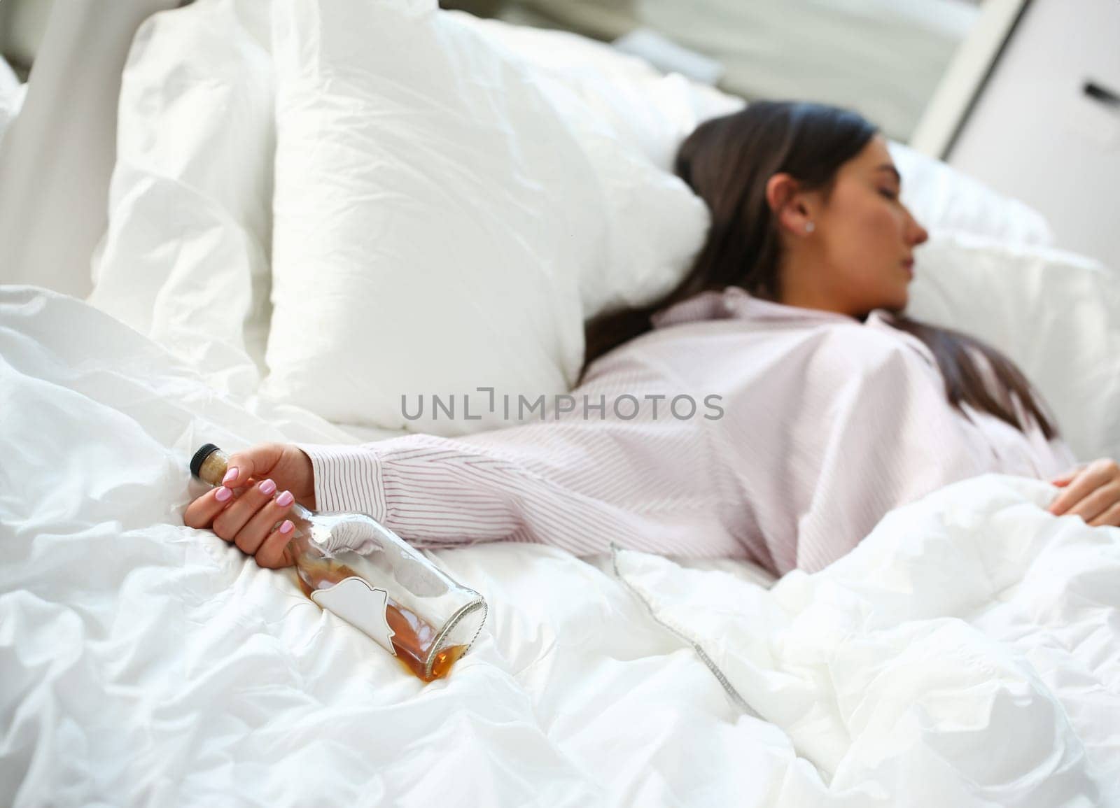 Young woman lying in bed deadly drunken holding near-empty bottle of booze. Female intoxicated with alcohol after tough night party. Alcoholism habitual drunkenness pernicious habit concept