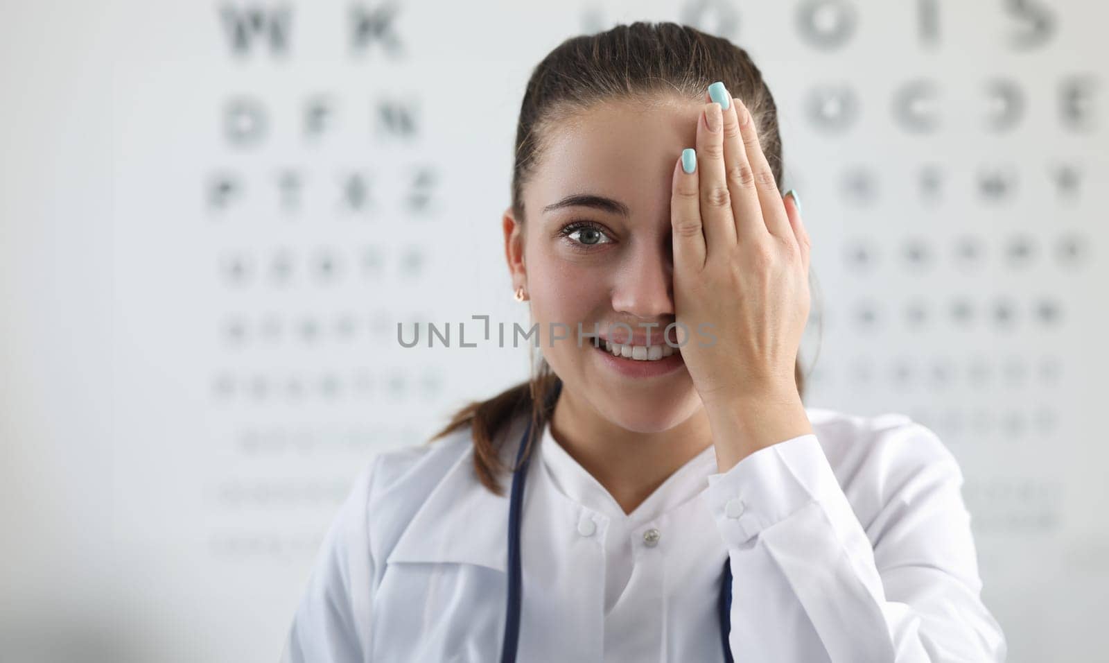 Portrait of lady oculist smiling and looking at camera with happiness. Cheerful doctor standing in clinic office and covering left eye with tender hand. Vision test concept