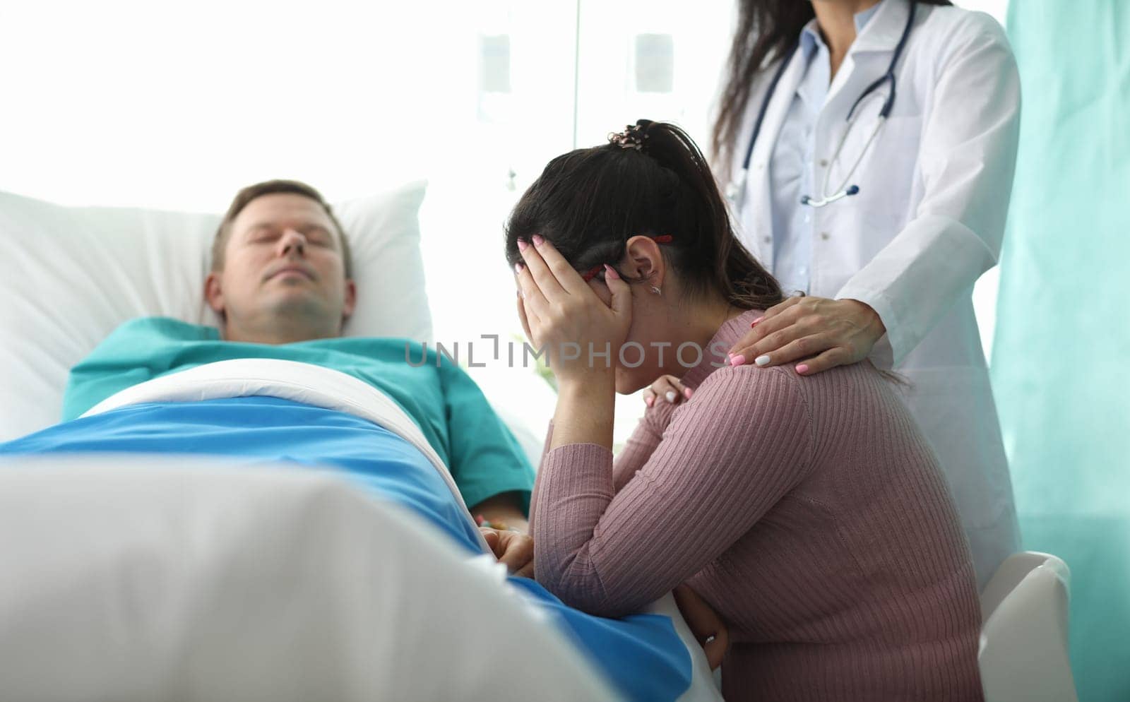 Portrait of beautiful doctor calming upset shocked wife. Ill man lying in hospital ward. Sad woman holding head by hands and crying. Healthcare and treatment concept