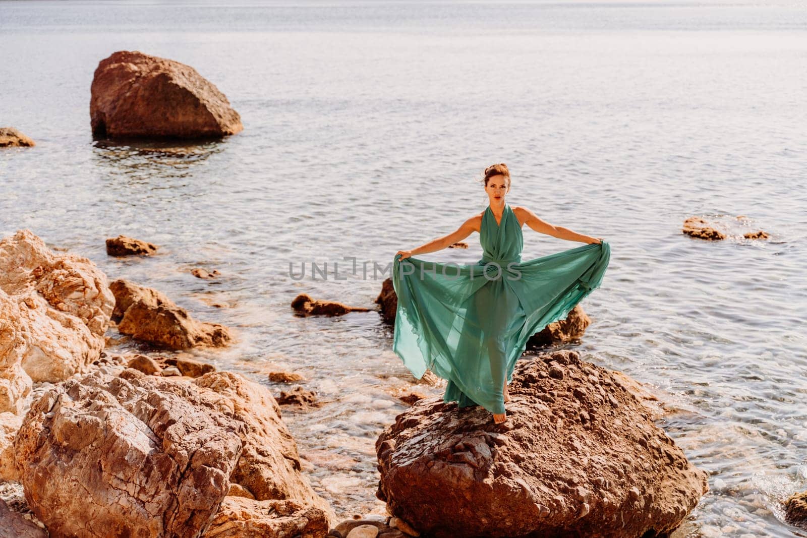 Woman green dress sea. Female dancer in a long mint dress posing on a beach with rocks on sunny day. Girl on the nature on blue sky background