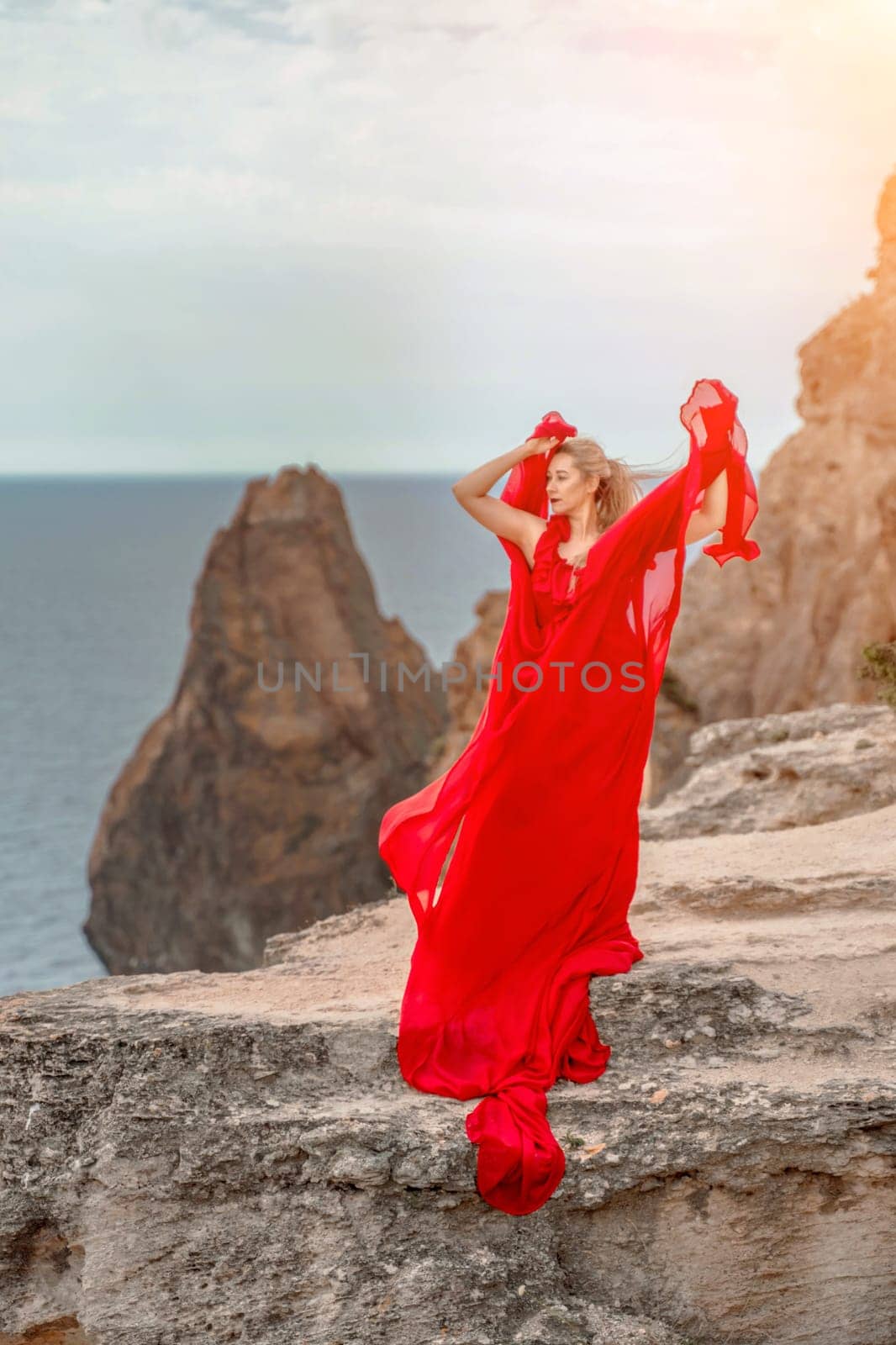 woman red silk dress stands by the ocean, with mountains in the background, as her dress sways in the breeze. by Matiunina