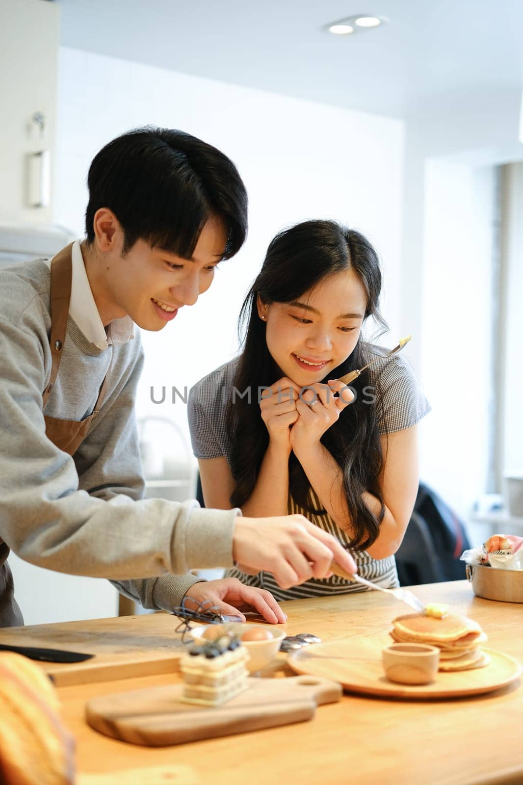 Smiling man putting butter on the hot pancakes, preparing breakfast for his girlfriend in cozy kitchen..