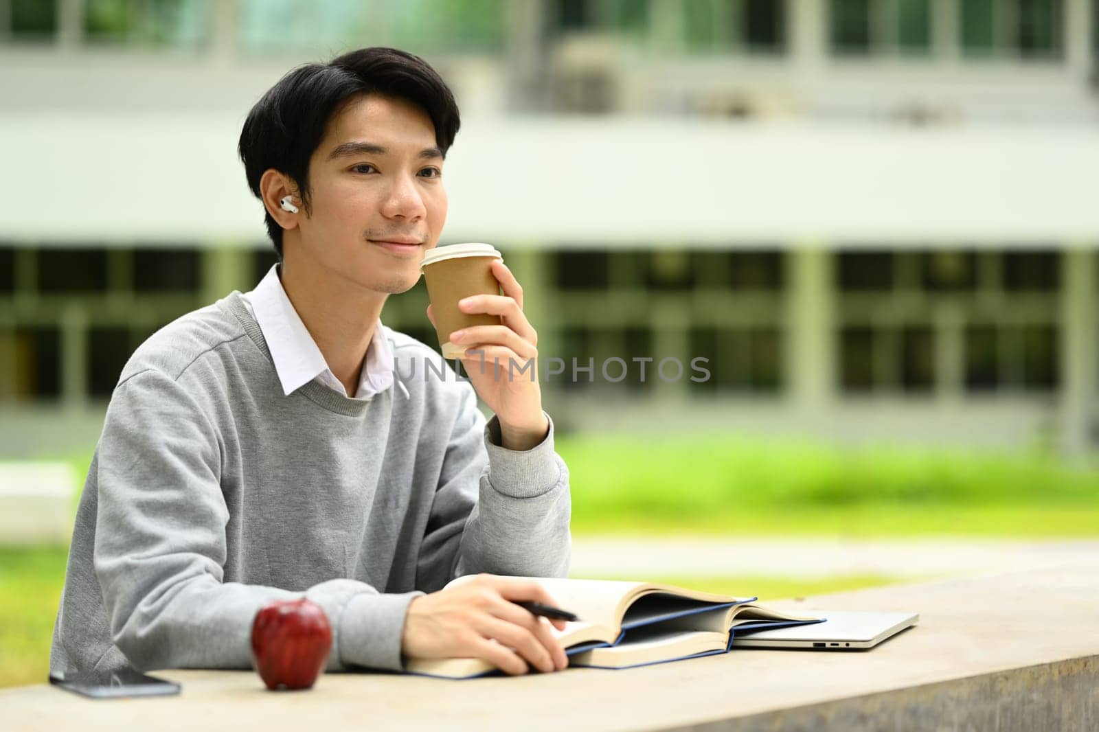 Relaxed man holding paper cup of coffee and looking away, sitting at table in front of university building.