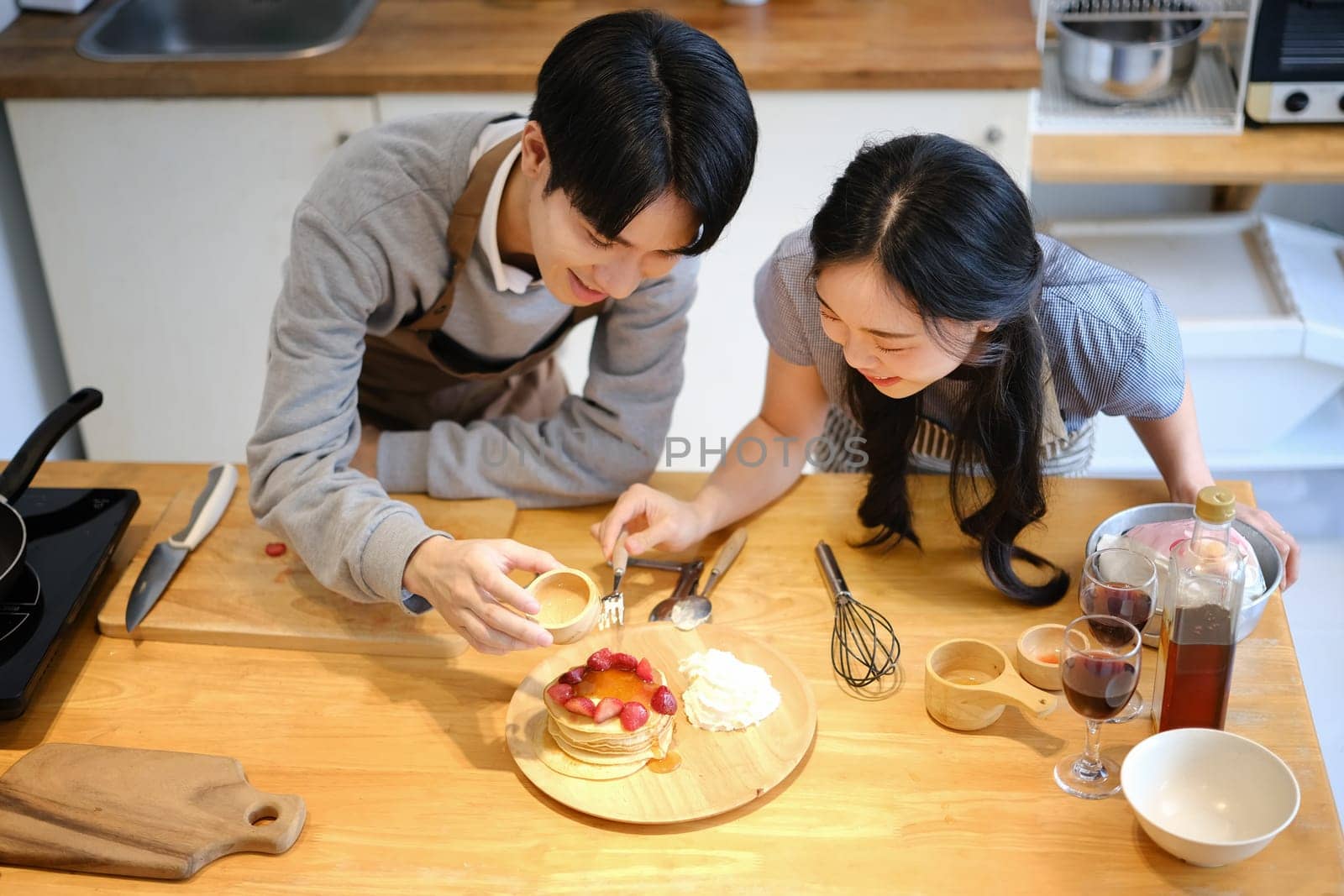 Joyful young couple putting honey on delicious pancakes with strawberries, preparing breakfast in cozy kitchen by prathanchorruangsak