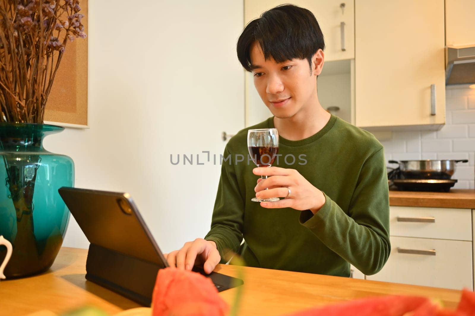 Smiling asian man in casual clothes holding glass of wine and using digital tablet on kitchen worktop.