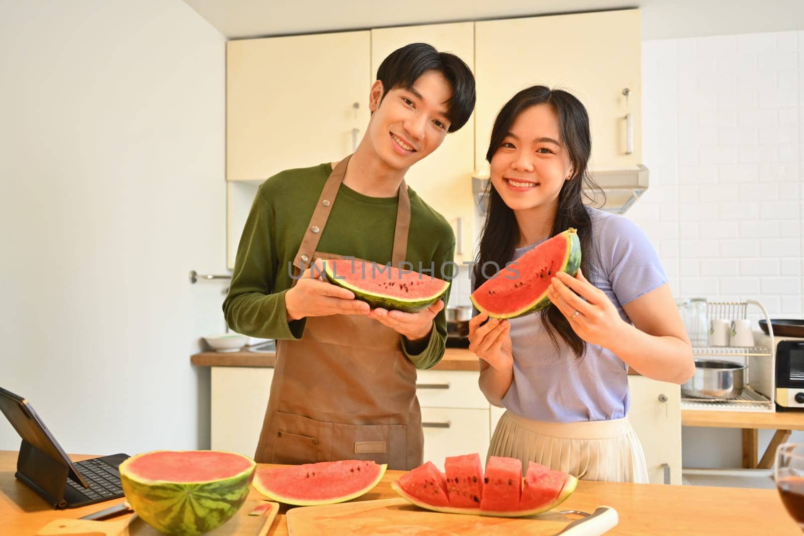 Happy young couple holding and eating slices of watermelon in home kitchen. Family love and casual lifestyle by prathanchorruangsak