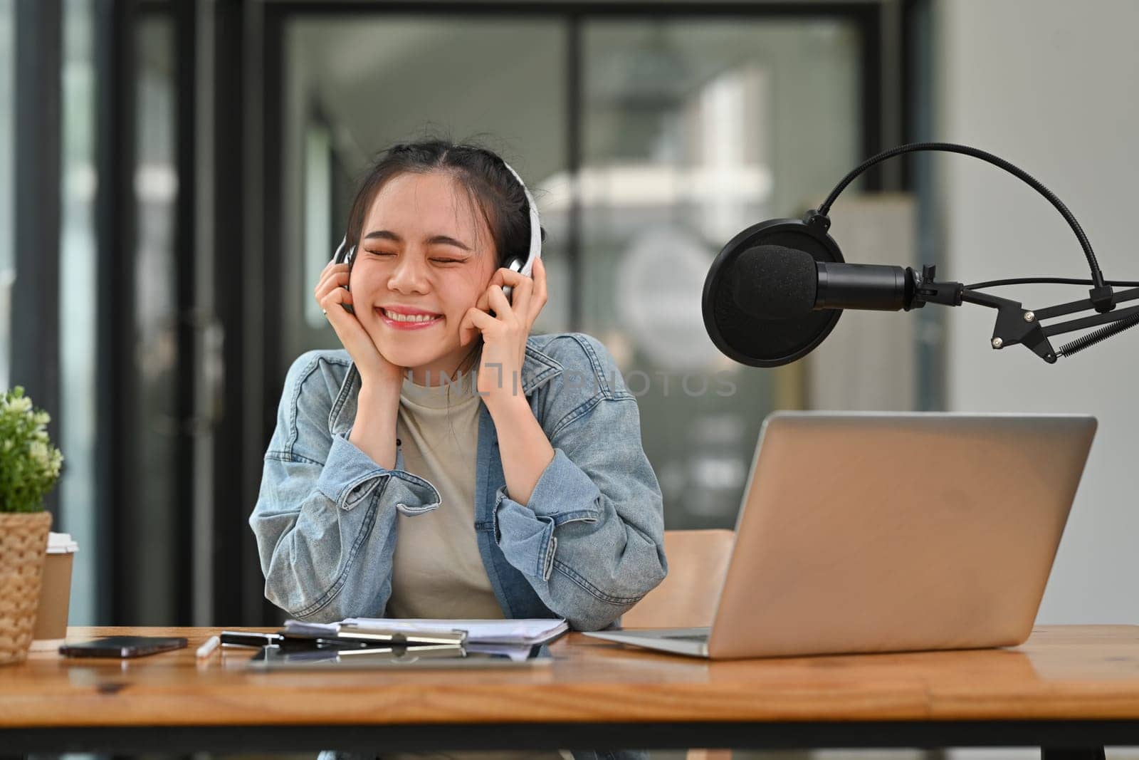 Cheerful Asian female radio host wearing headphone recording podcast at home studio.