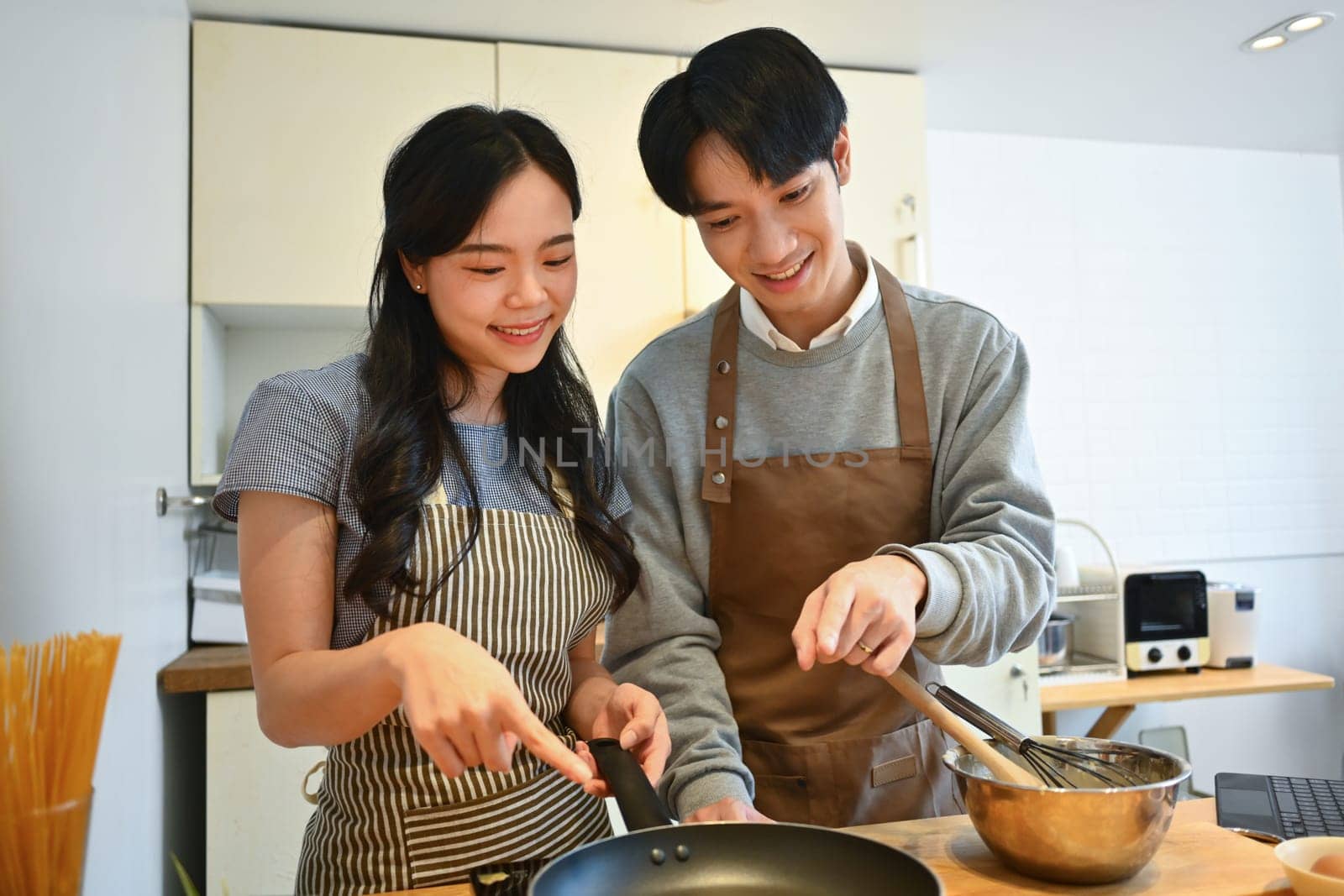 Happy young Asian couple on aprons making pancakes for breakfast, spending a weekend morning together by prathanchorruangsak