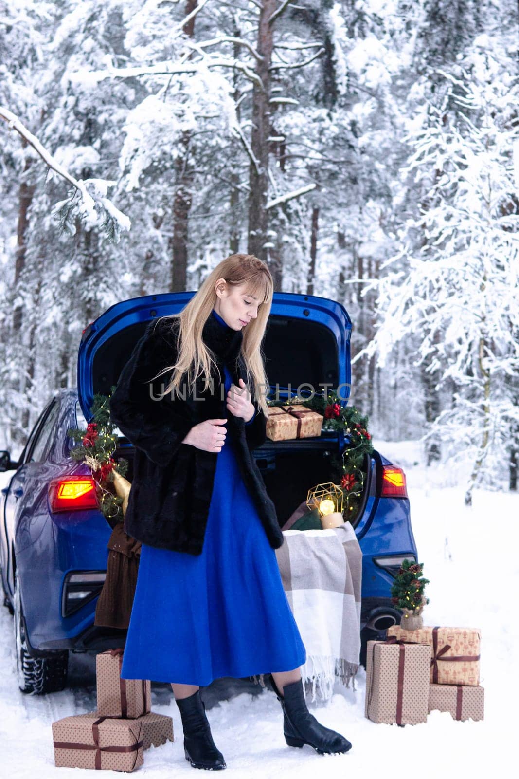 Woman in winter snowy forest in blue dress next to blue car decorated with Christmas decor. Christmas and winter holidays concept