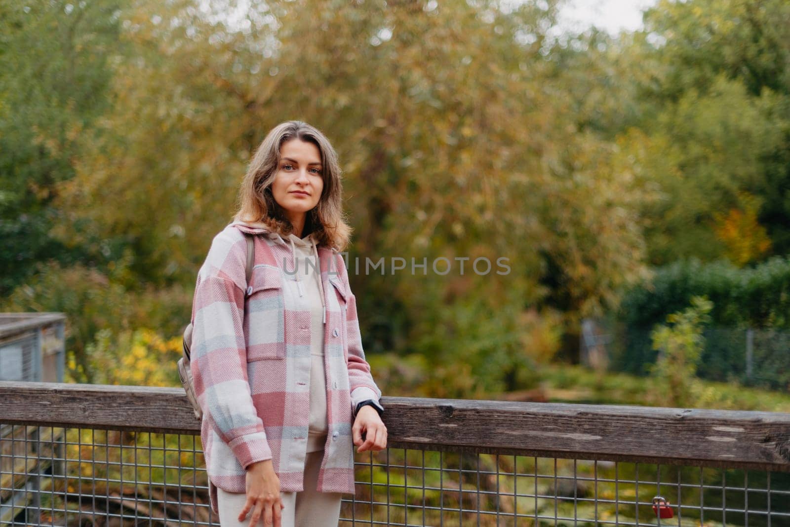 Portrait of cute young woman in casual wear in autumn, standing on bridge against background of an autumn Park and river. Pretty female walking in Park in golden fall. Copy space. smiling girl in the park standing on wooden bridge and looking at the camera in autumn season