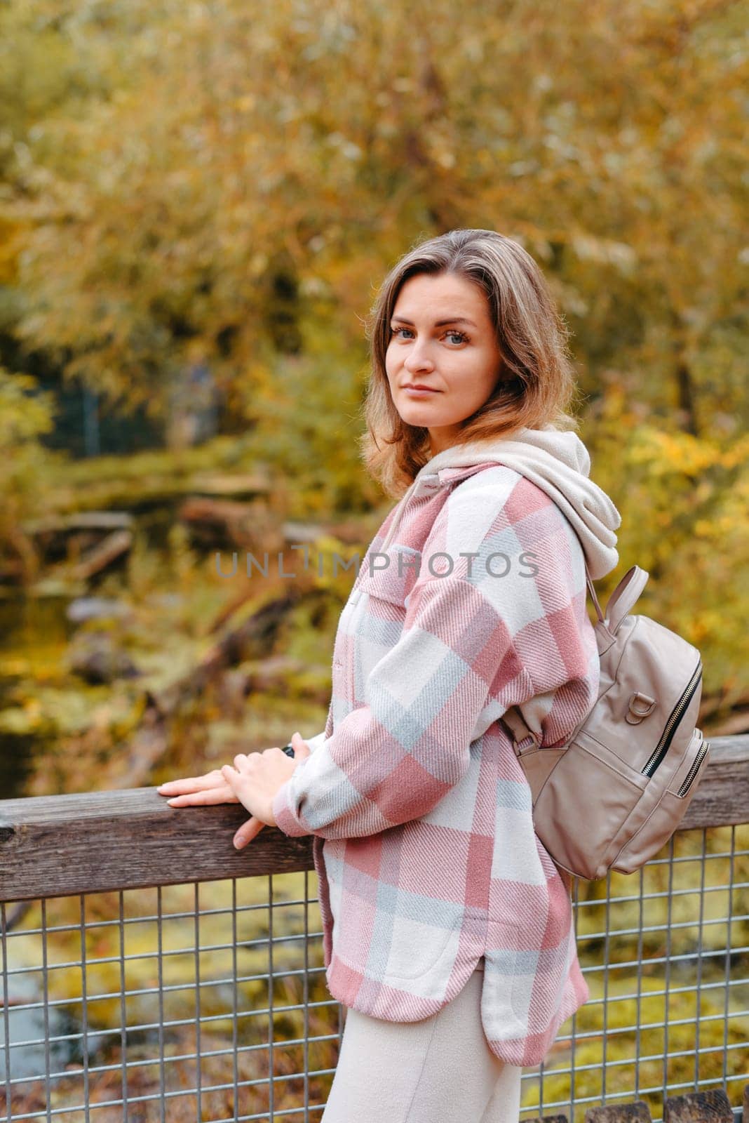 Portrait Of Cute Young Woman In Casual Wear In Autumn, Standing On Bridge Against Background Of An Autumn Park And River. Pretty Female Walking In Park In Golden Fall. Copy Space. Smiling Girl In The Park Standing On Wooden Bridge And Looking At The Camera In Autumn Season by Andrii_Ko