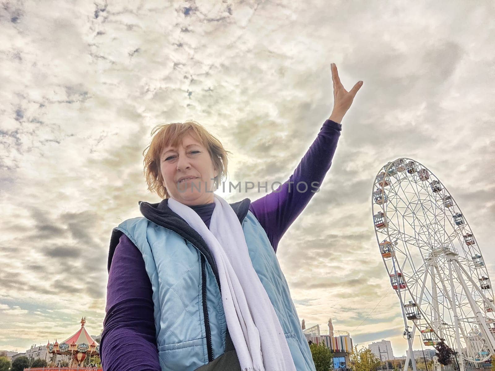 Relaxed mature caucasian female making selfie outside near big Ferris wheel. Positive senior woman against city street background. Adult middle agged girl Blogger taking photo for internet by keleny