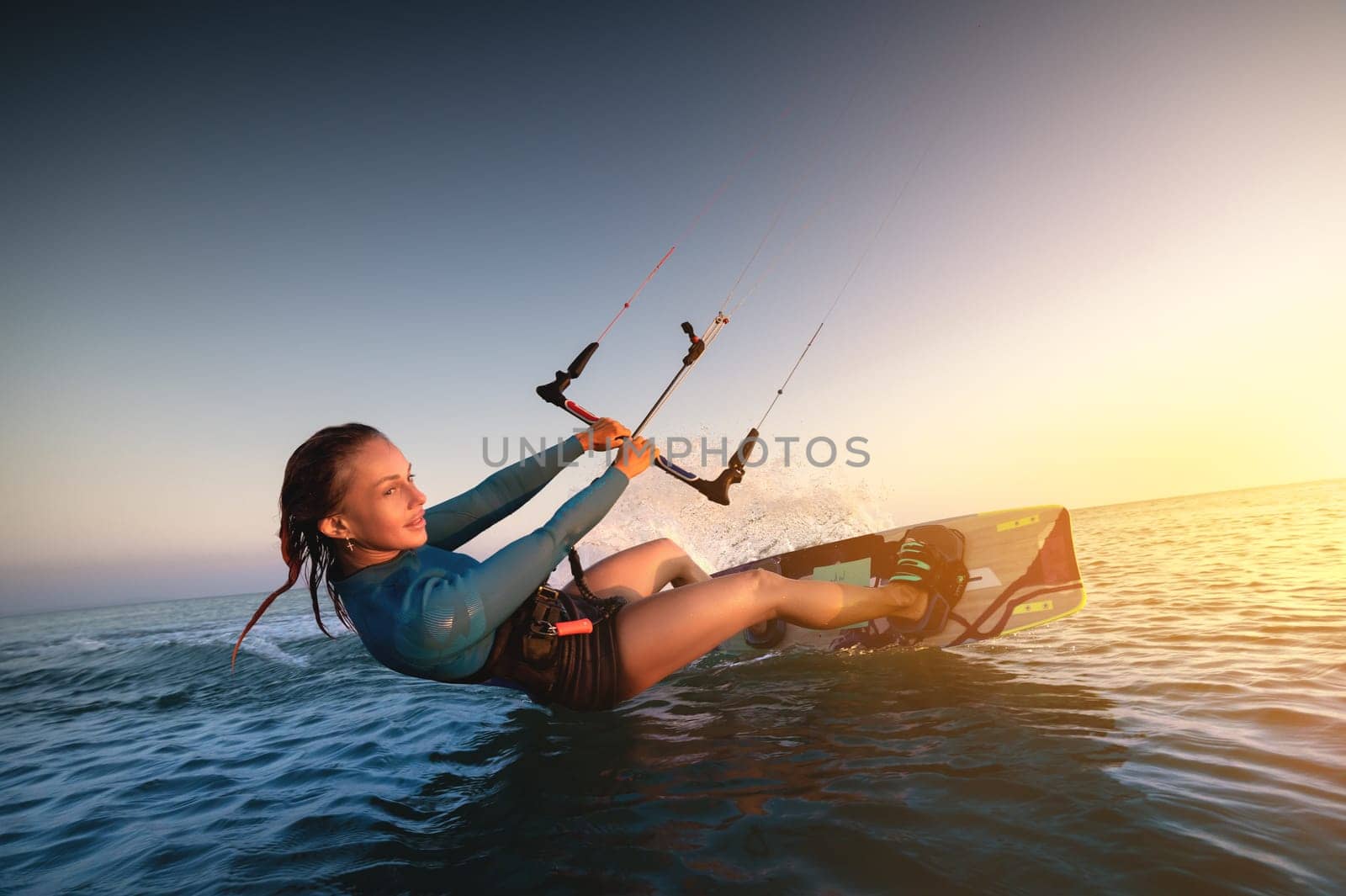 Girl kitesurfing in a sexy hydrosuit with a kite in the sky on board in the blue sea, riding on the water waves. Recreational activities, water sports, activities, hobbies and entertainment in the summer. Kiteboarding.