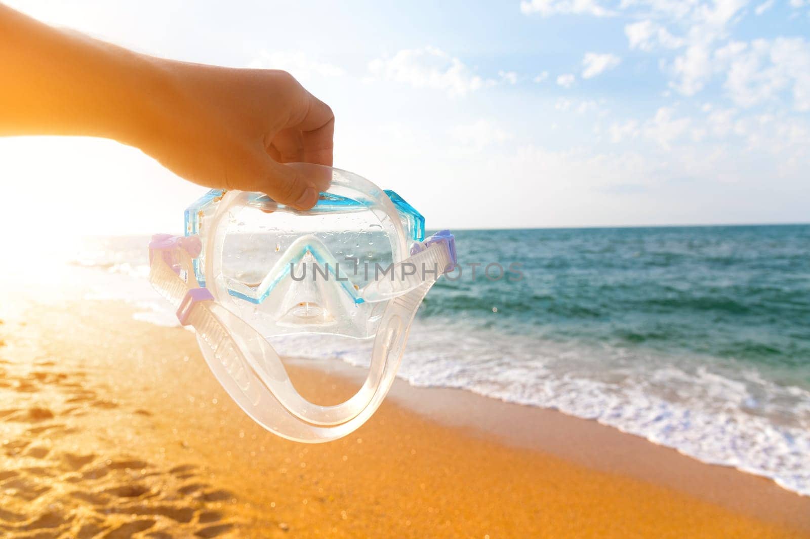 A hand holds snorkeling goggles against the background of the beach and sky. A man's hand holds a pipe over a sandy beach on a sunny day. Water sports. Diving. Travel and vacation concept
