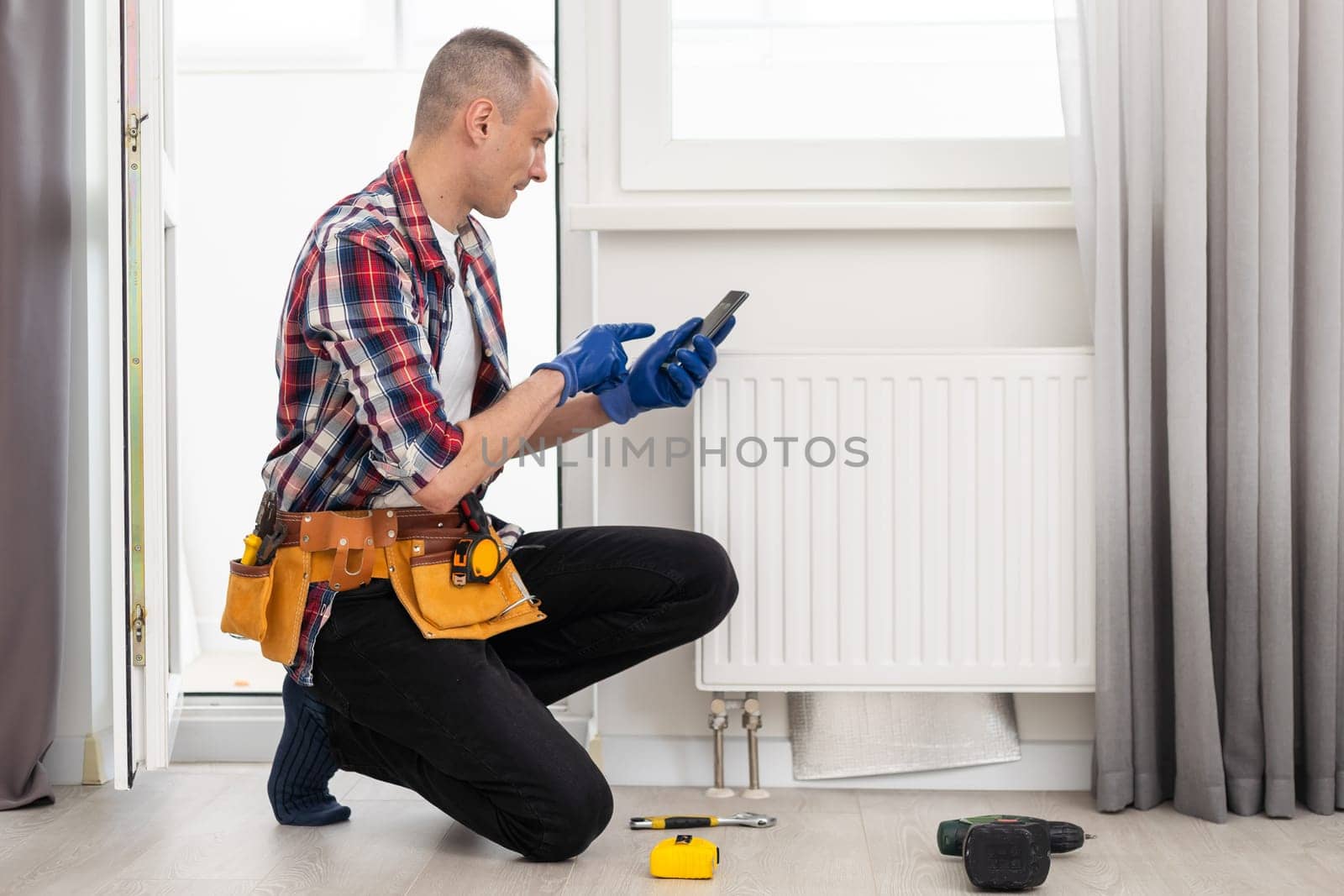 Man in work overalls using wrench while installing heating radiator in room. Young plumber installing heating system in apartment. Concept of radiator installation, plumbing works and home renovation by Andelov13