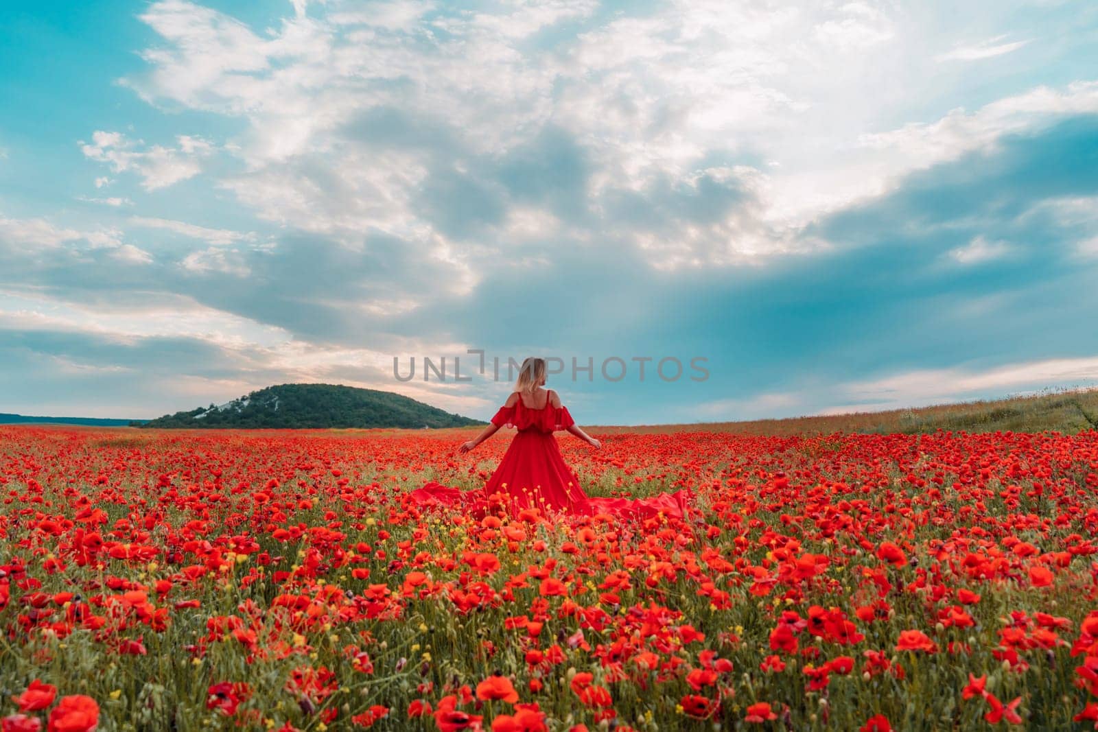 Happy woman in a long red dress in a beautiful large poppy field. Woman stands with her back in a long red dress, posing on a large field of red poppies.