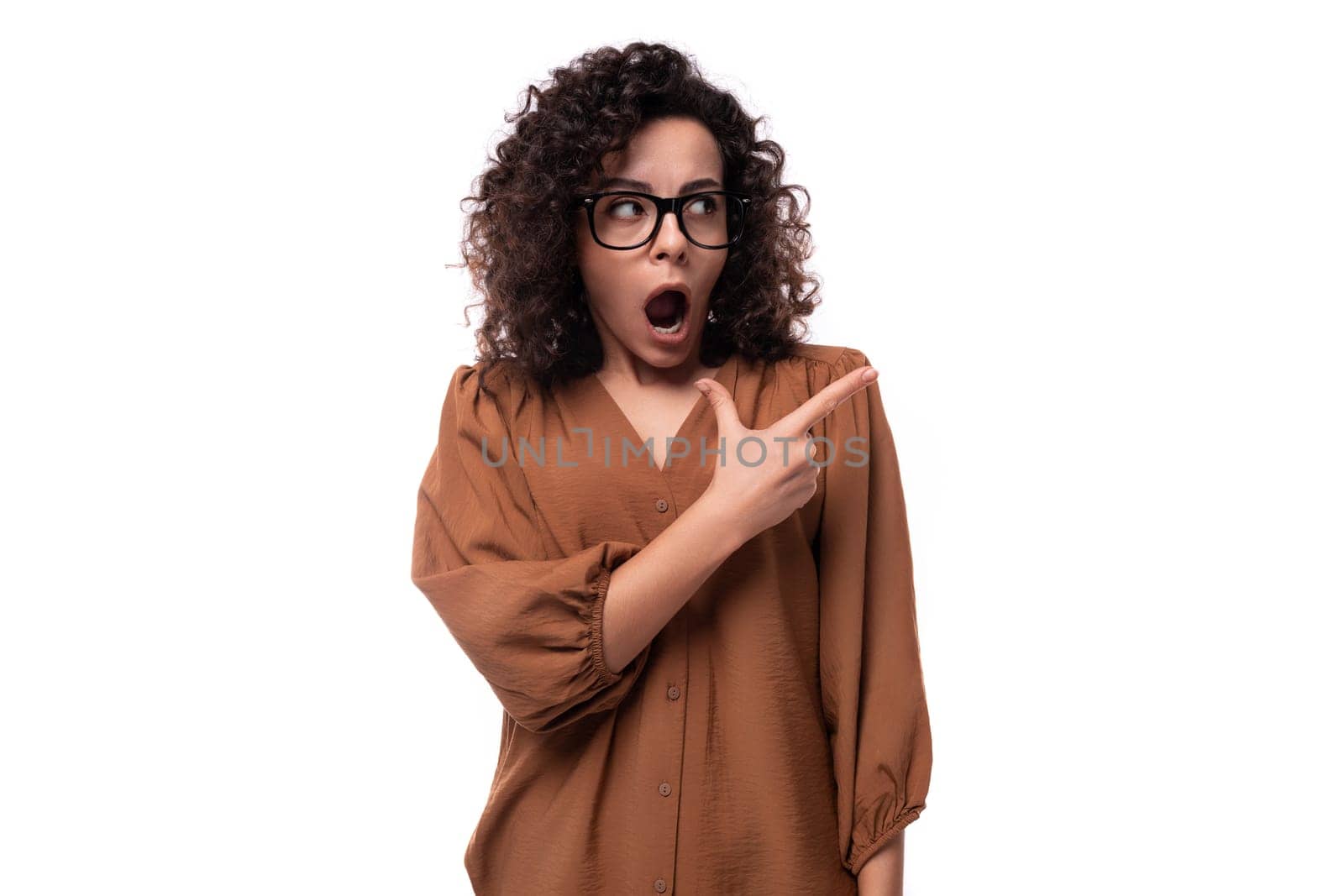 young curly businesswoman dressed in a brown shirt points her hand to the side.