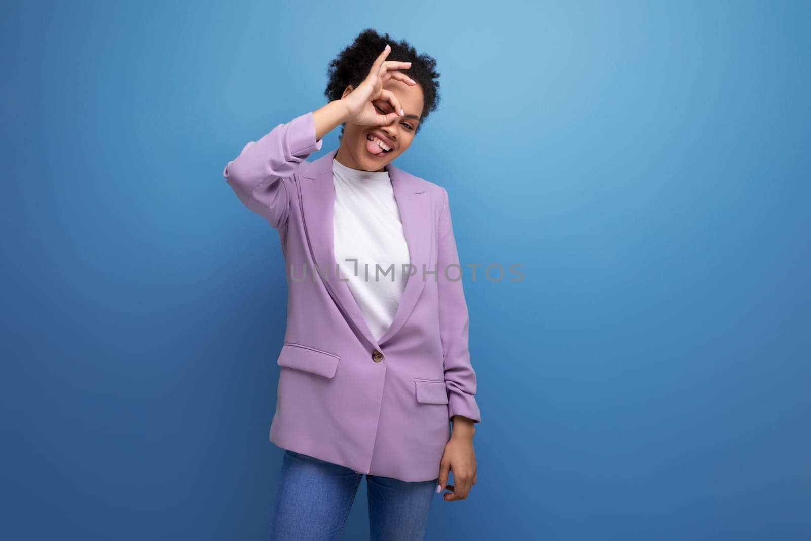 young cute hispanic business woman with curly hair in a lilac jacket smiling on a blue background with copy space.