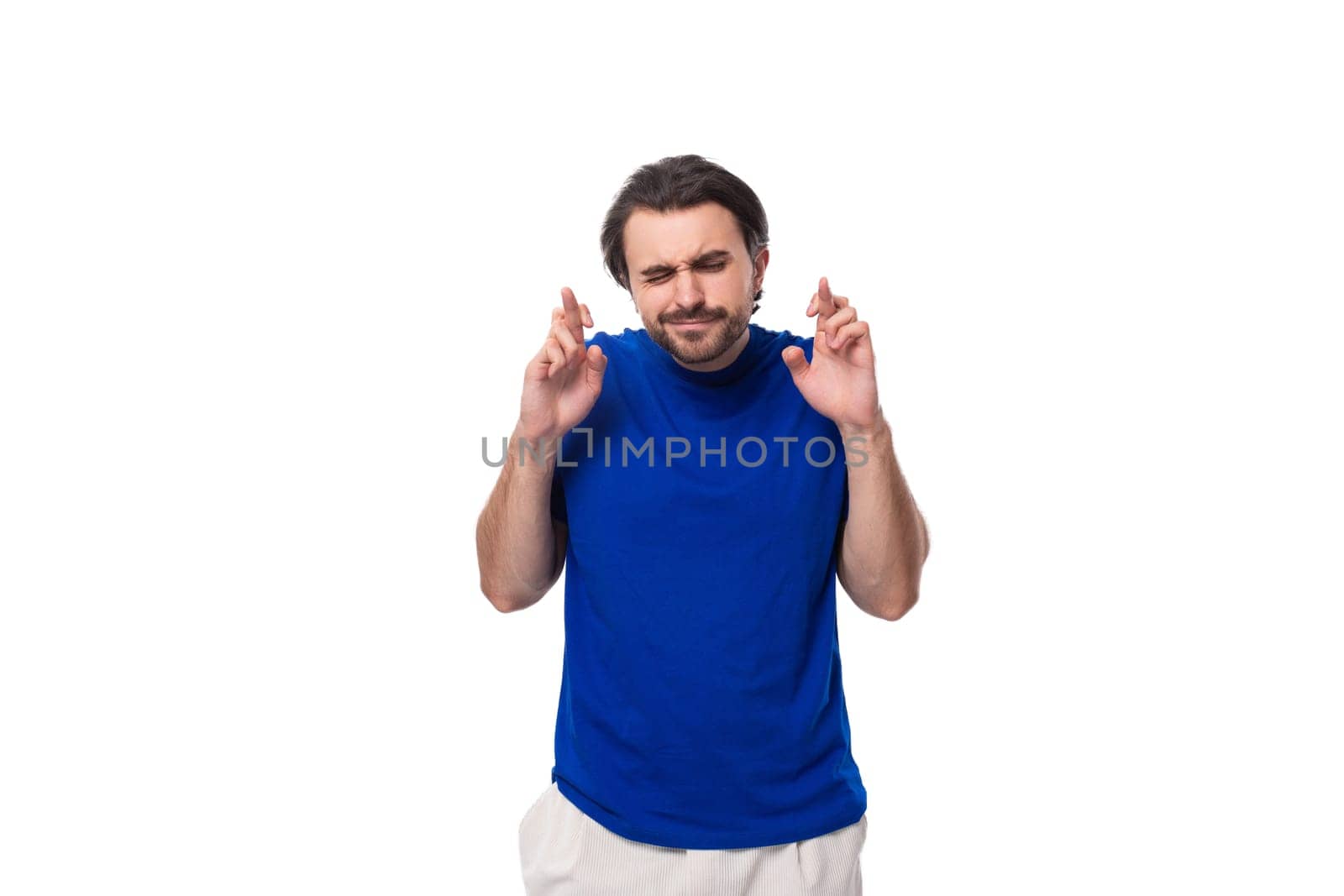 young brunette man with a beard dressed in a blue t-shirt crossed his fingers in hope on a white background.