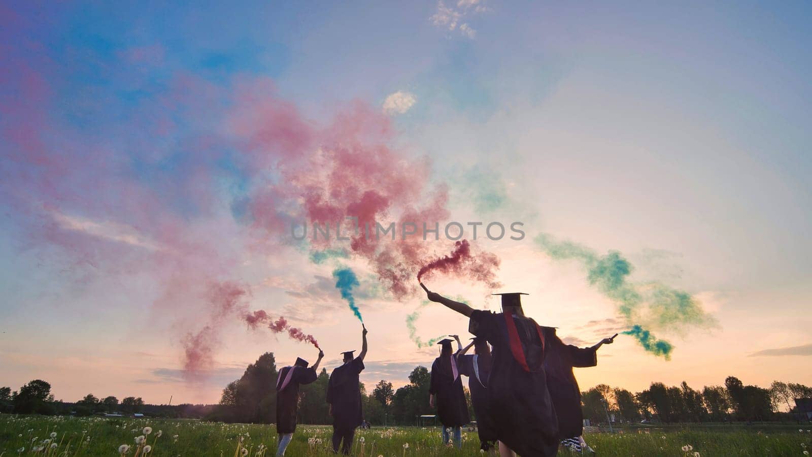 Graduates in costume walk with a smoky multi-colored smoke at sunset.