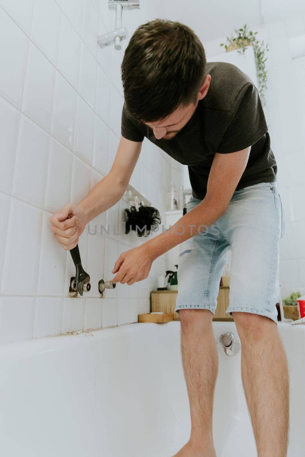 One young brunette Caucasian man tightens the faucet nut with both hands using an adjustable wrench in the bathroom sticking out of the wall, standing in the bathroom during the day, close-up side view.Step by step.