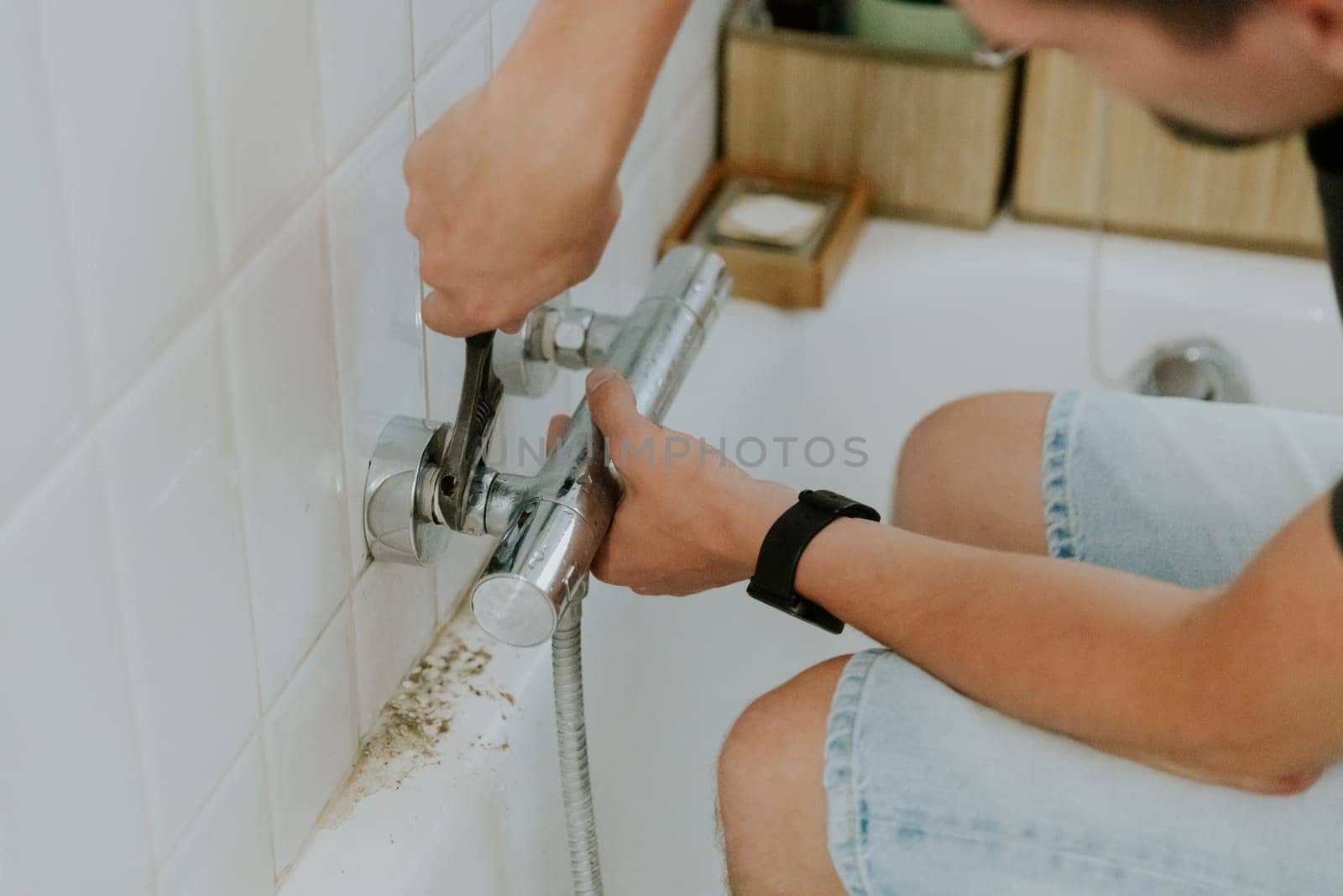 One young brunette Caucasian guy manually installs a faucet into the walls using an adjustable wrench while sitting on the edge of the bathtub, close-up side view. Step by step.