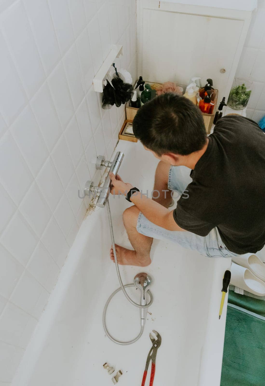 One young Caucasian brunette removes an unscrewed faucet in the wall using his hands, sitting on the edge of the bathtub, top view. Step by step.