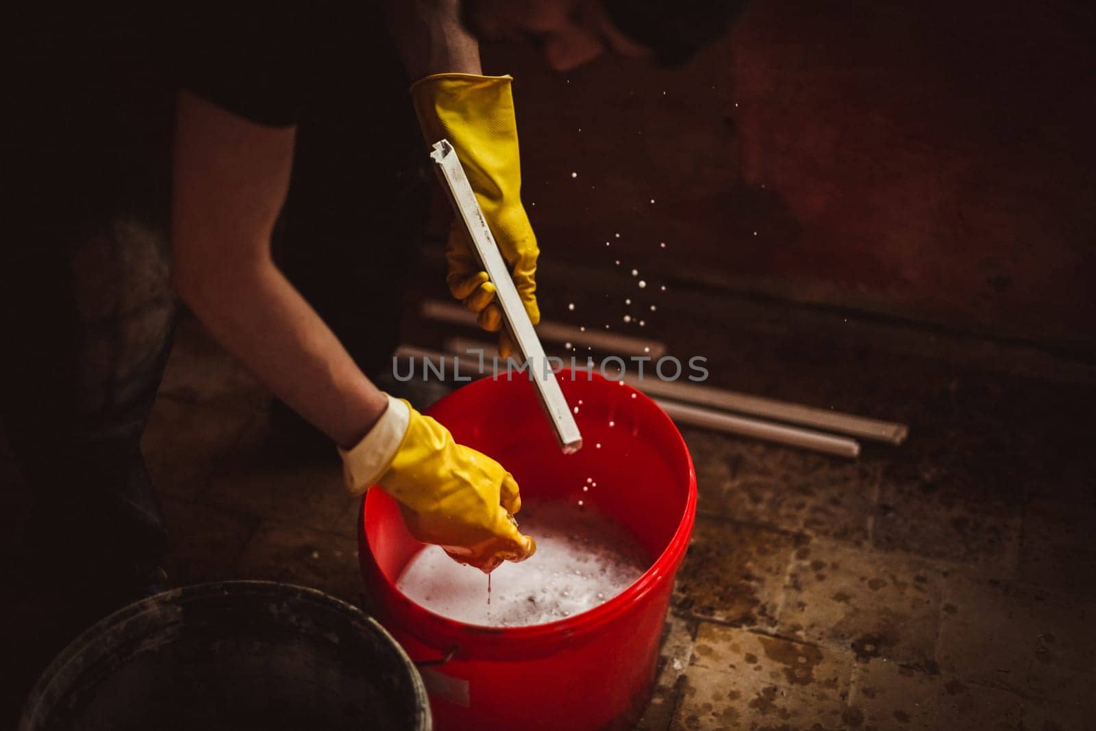 A young Caucasian man in black clothes with a cap on his head and yellow rubber gloves washes window frames in a bucket of soapy water, squeezing with a sponge, standing bent over in a room where repairs are underway, close-up side view.