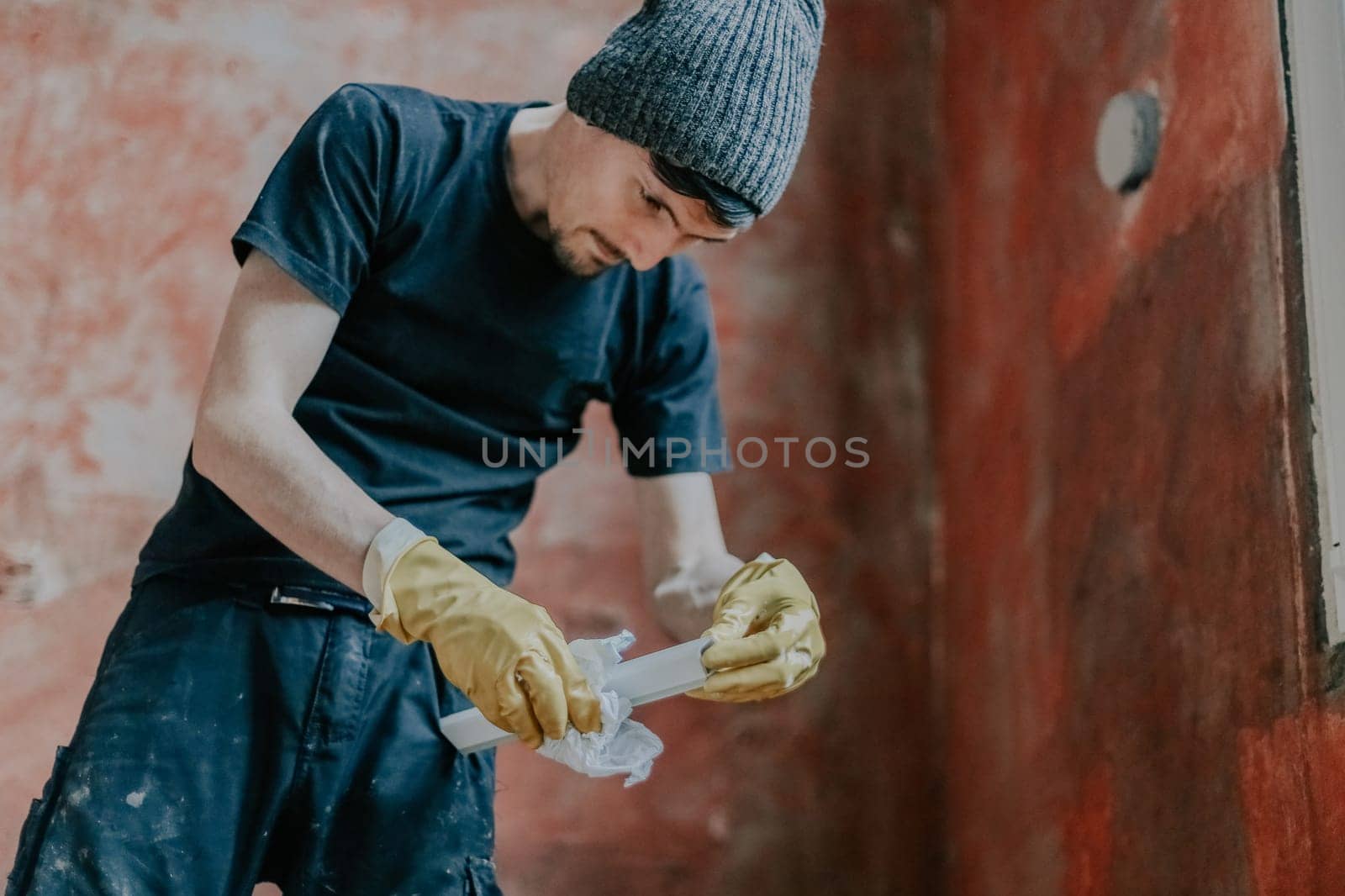 A young Caucasian man in black clothes, with a cap on his head and wearing yellow rubber gloves washing window frames, standing in a room against a red wall, close-up view from below.