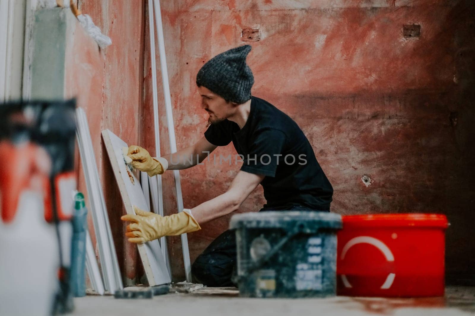 One young Caucasian man in black clothes and yellow rubber gloves washes the plastic part of the window frames with soapy water and a sponge, sitting on his knees in the corner of the room where repairs are underway, close-up view from below.