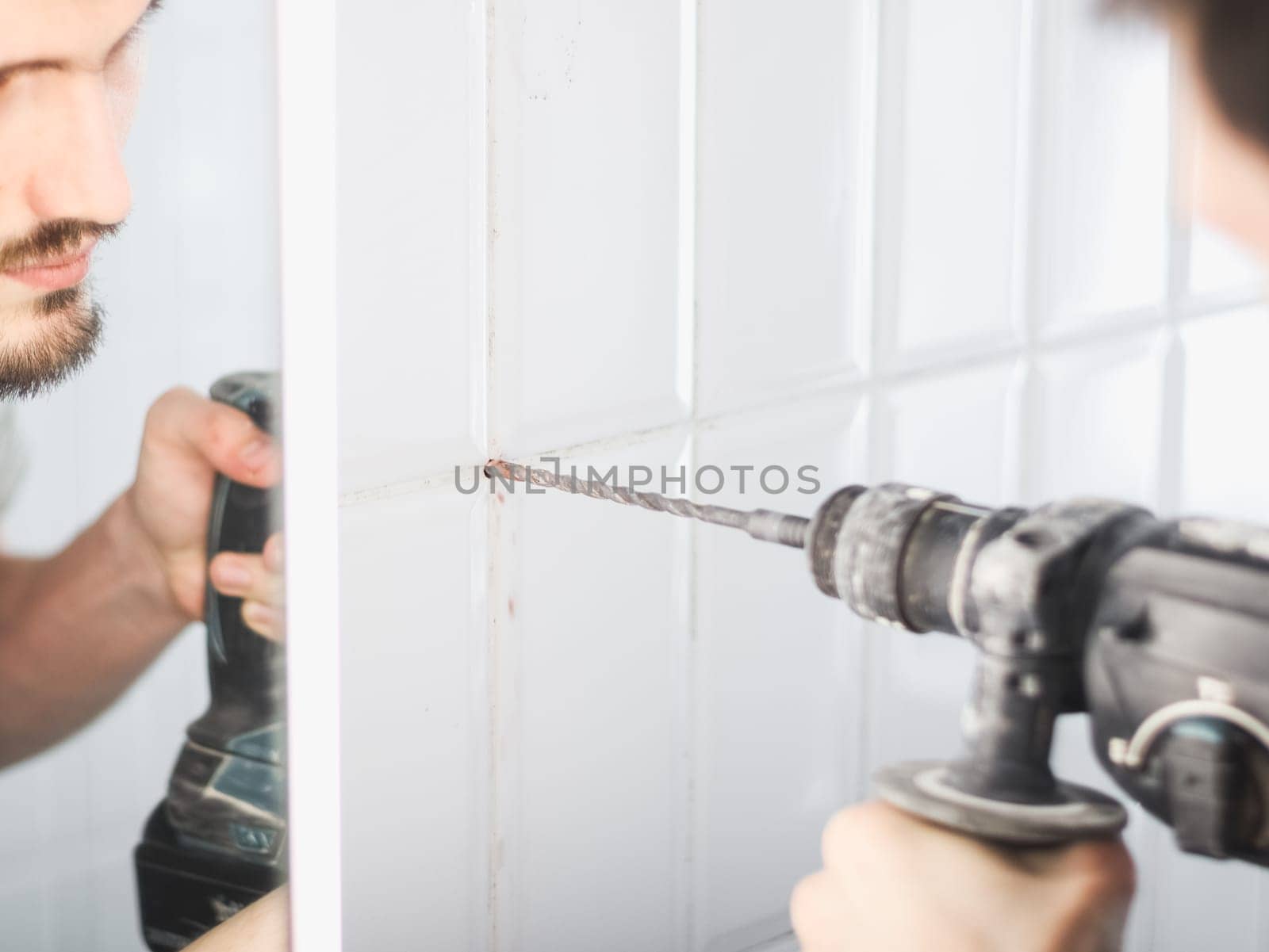 One young Caucasian man drills a hole between the seams of a tiled wall using a drill in the bathroom, reflected in the mirror of a wall cabinet, close-up view from below.