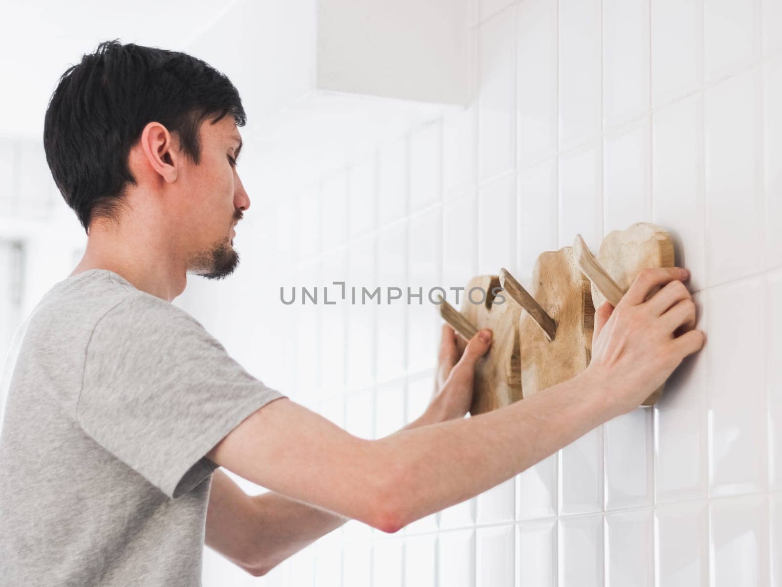 One young Caucasian man stands sideways and hangs a creative wooden towel rack on a white tiled wall in the bathroom, close-up side view.