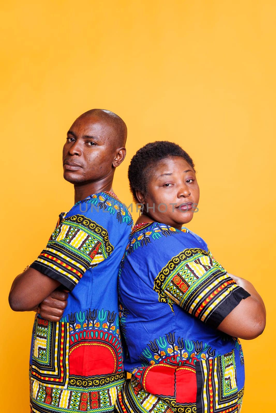 Serious black couple dressed in ethnic clothes posing with arms crossed studio portrait. Confident man and woman standing back to back with folded hands and looking at camera