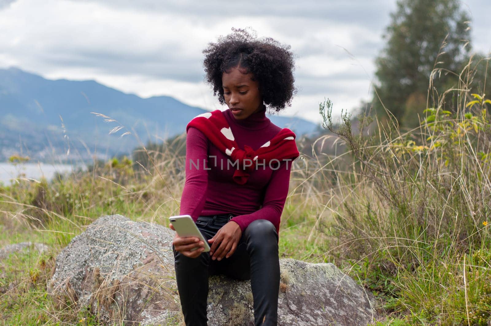 afro girl checking her cell phone before a live broadcast sitting on a mountain rock by Raulmartin
