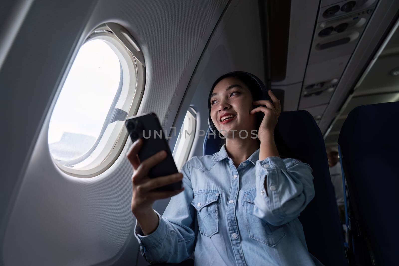 Asian people female person onboard, airplane window, using mobile and headphone while on the plane.