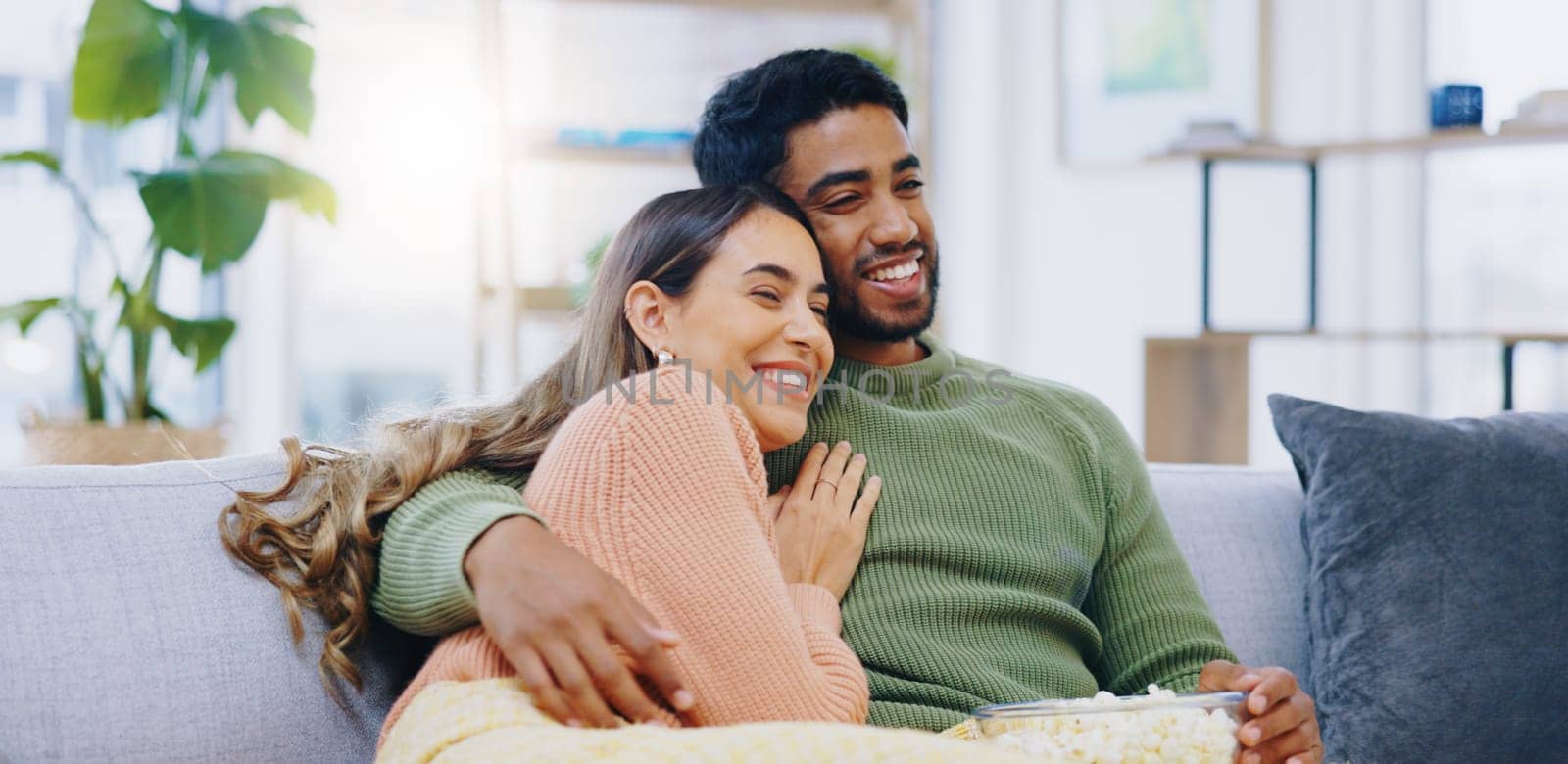 Love, hug and couple on watching tv on a sofa with popcorn for movie, film or streaming show at home. Relax, television and people embrace in a living room with cinema snack for comedy or series.