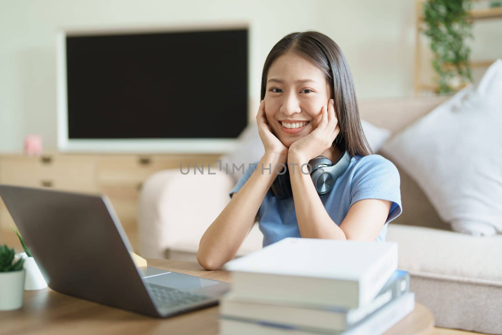 Portrait of young beautiful Asian woman showing smiling face during early morning online class with books, headphones and computer as study materials at home by Manastrong