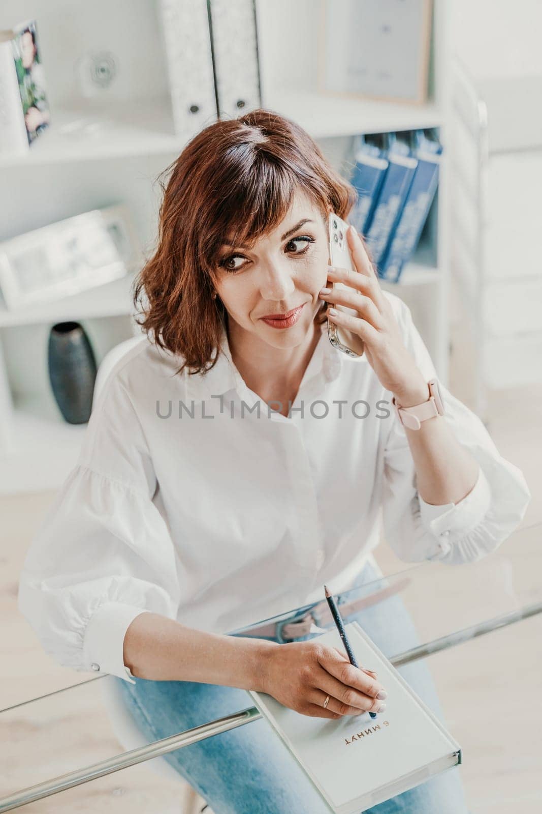 Woman in the office at the computer. She is dressed in a white shirt and blue jeans sitting at a table on which is a laptop and a notepad. Works in the office, bright workplace