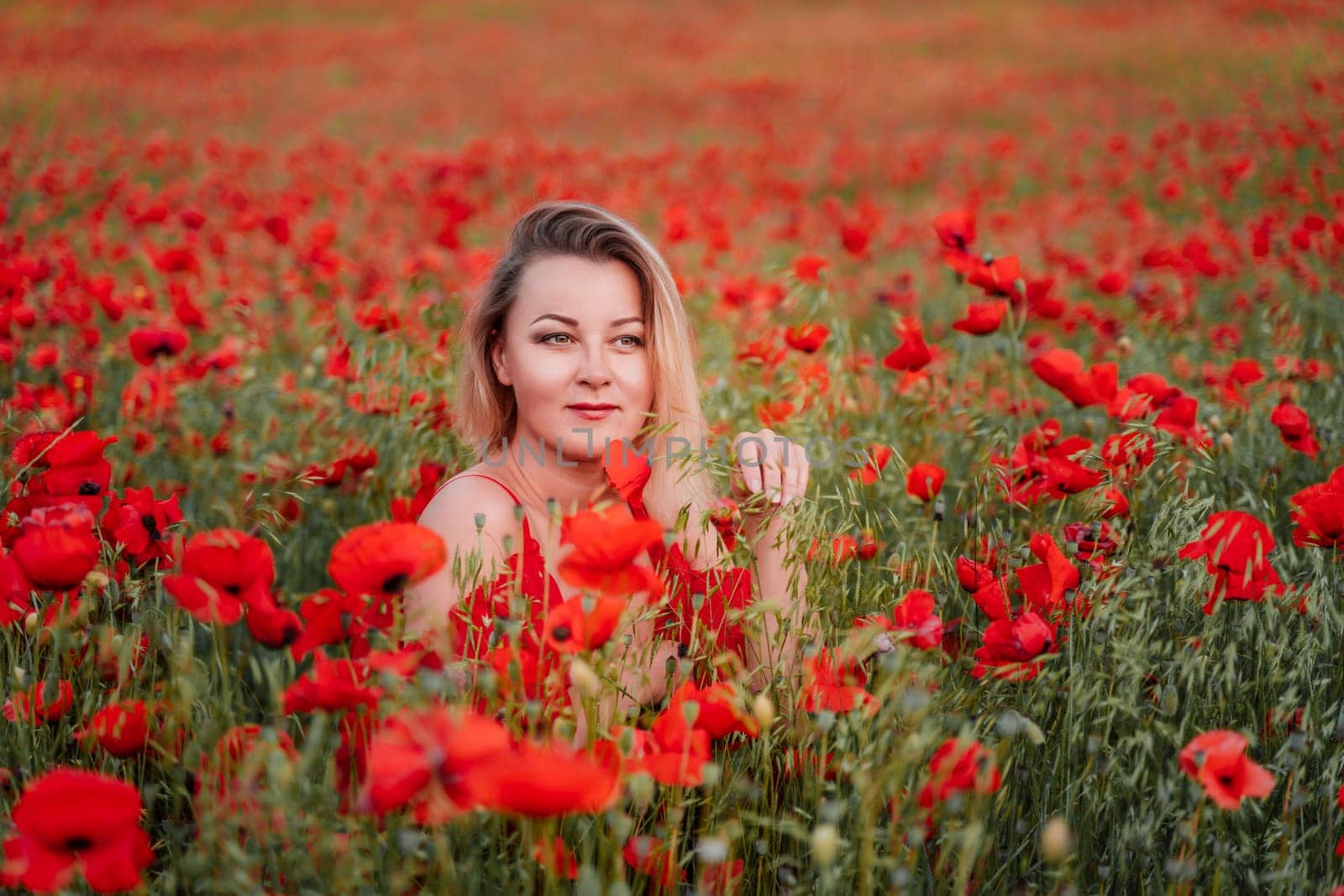 Happy woman in a red dress in a beautiful large poppy field. Blond sits in a red dress, posing on a large field of red poppies.