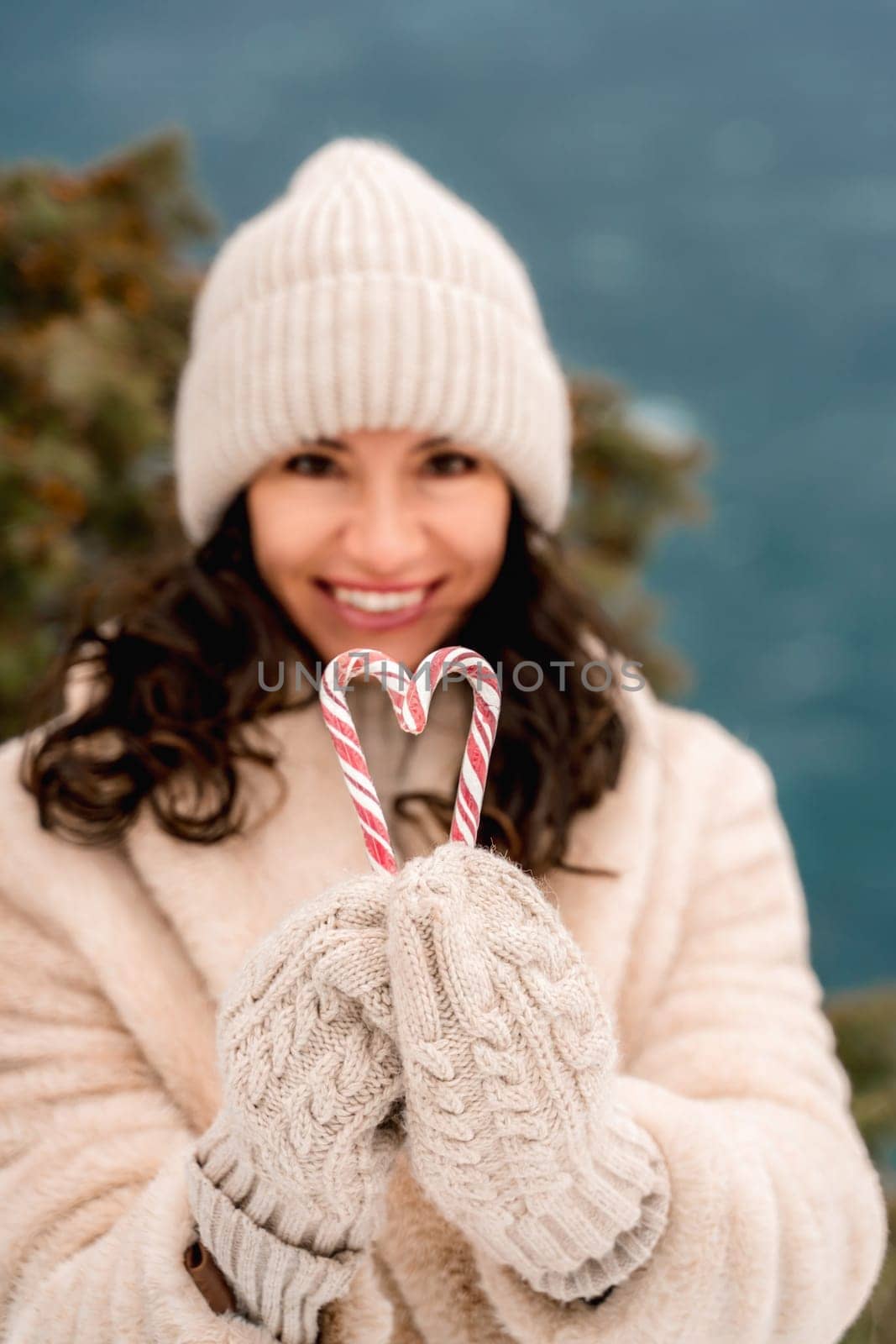 Woman candy sea. Smiling woman in knitted hat, mittens and beige coat holding lollipops candy canes in her hands in shape of heart against the backdrop of the sea