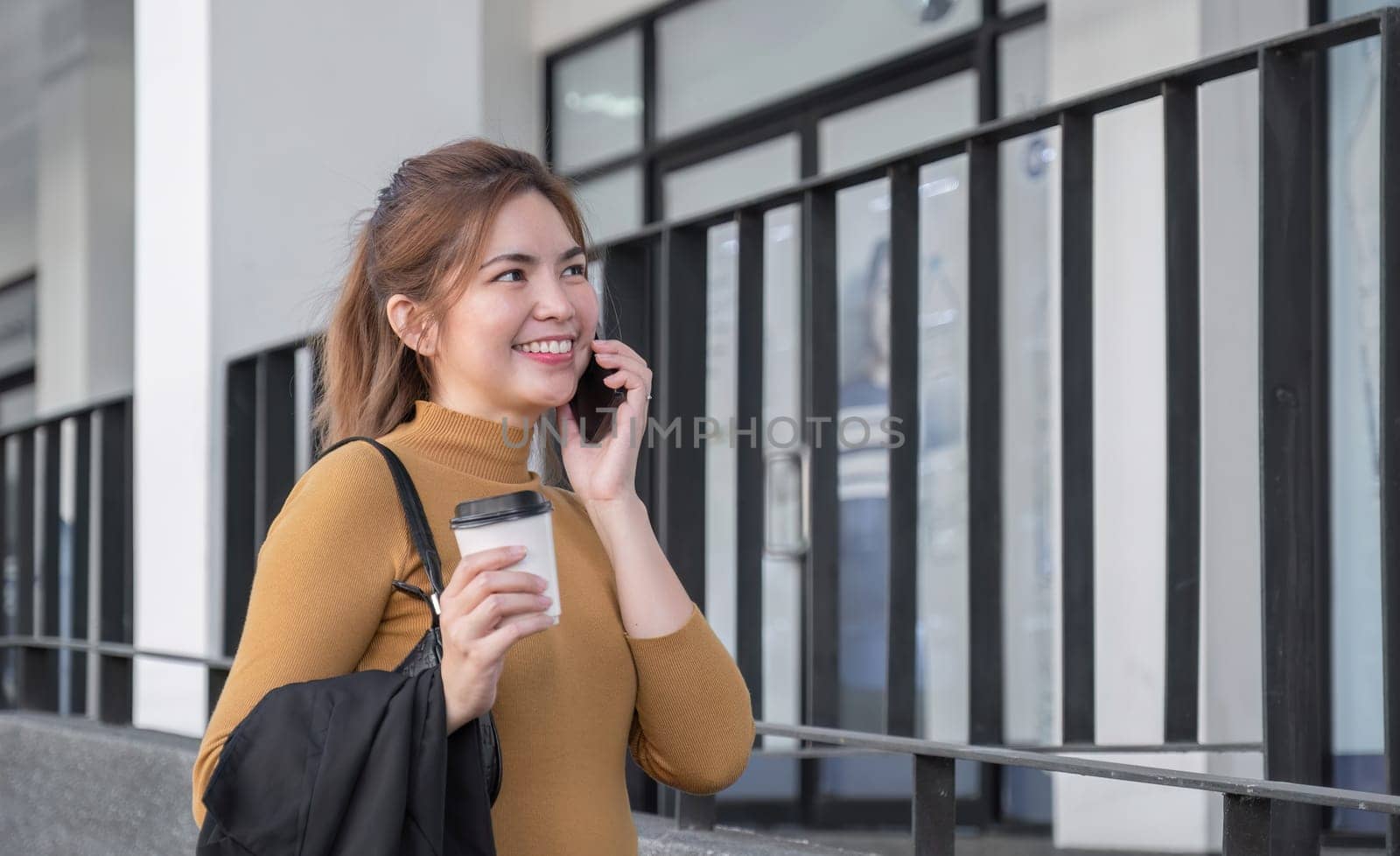 Asian businesswoman smiling while talking on mobile phone and holding coffee cup outside the office. by wichayada