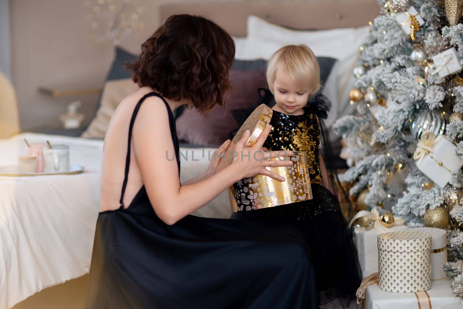 Mother daughter 2 years old Christmas tree. Both are dressed in black dresses, the mother holds the girl in her arms and both look at each other. The family celebrates Christmas