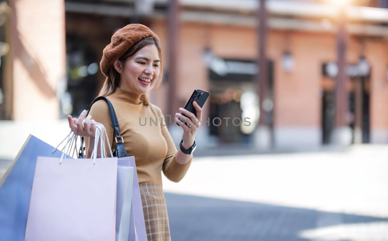 Beautiful Asian woman holding shopping bags, using smartphone and smiling while shopping on Black Friday outside the mall. by wichayada