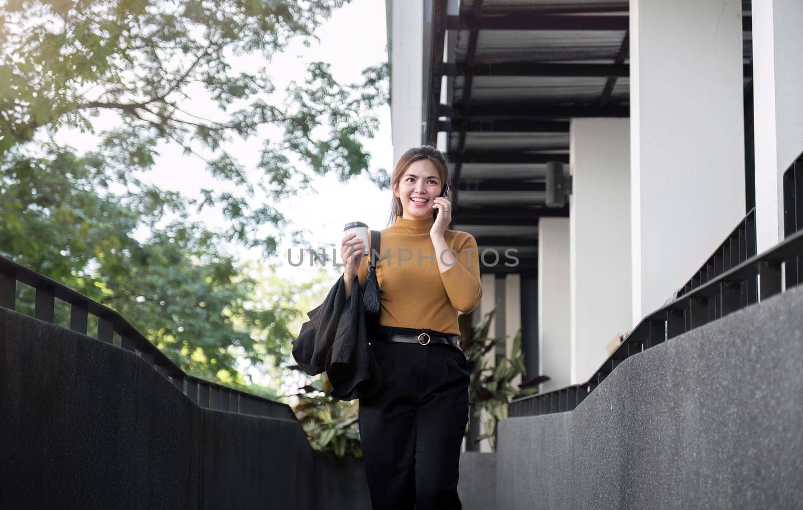 Asian businesswoman smiling while talking on mobile phone and holding coffee cup outside the office. by wichayada