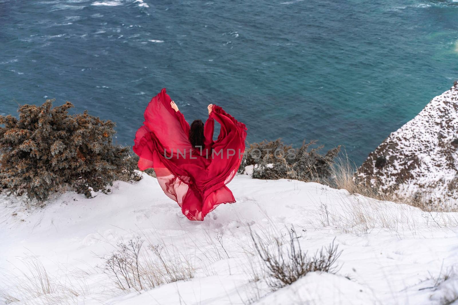 Woman red dress snow sea. Happy woman in a red dress in the snowy mountains by the emerald sea. The wind blows her clothes, posing against sea and snow background