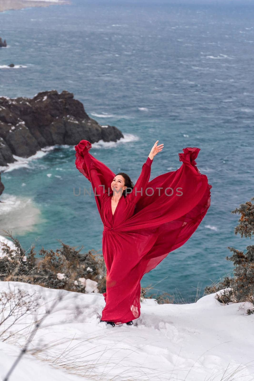 Woman red dress snow sea. Happy woman in a red dress in the snowy mountains by the emerald sea. The wind blows her clothes, posing against sea and snow background. by Matiunina